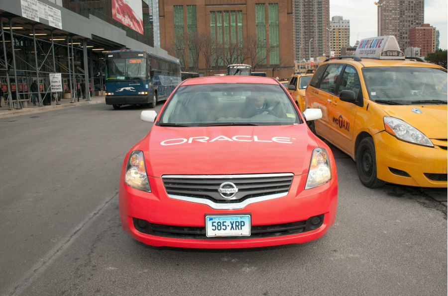Bild eines Autos, das auf einer Straße fährt, auf dem Oracle geschrieben ist. Auf der einen Seite ist ein Bus, auf der anderen ein Taxi.