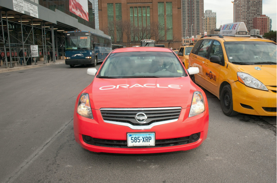Picture of a car driving on a road, with Oracle written on it. To one side is a bus, to the other a taxi.
